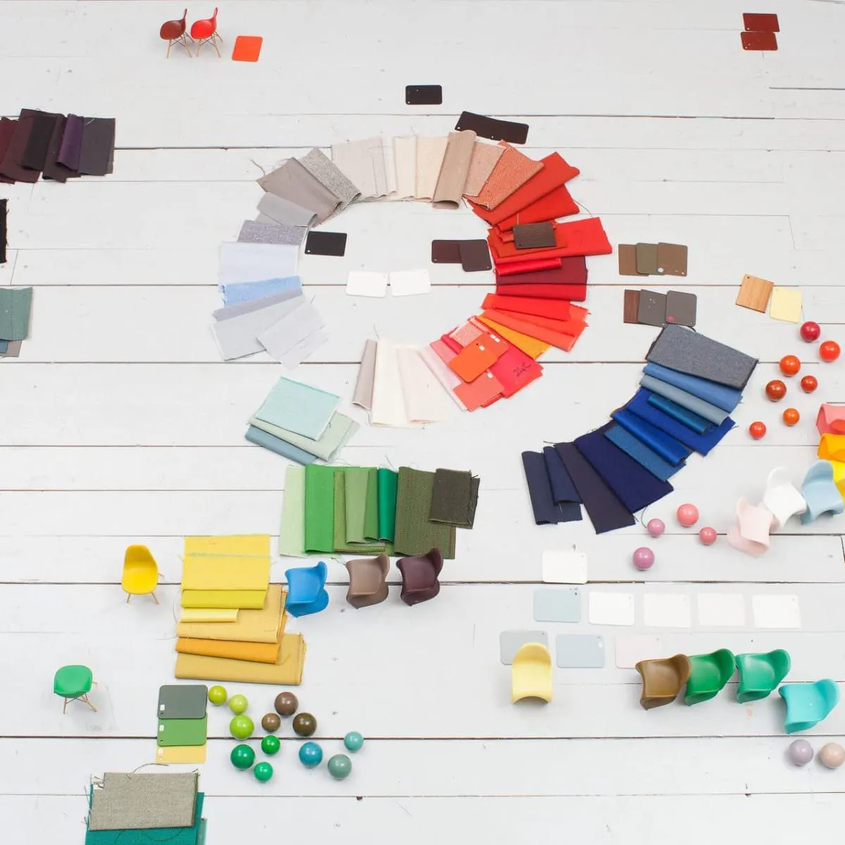 Vitra Colour & Material Library vista cenital: cientos de muestras textiles dispuestas en espiral cromática sobre suelo blanco, mini sillas Eames y Panton de colores distribuidas como referencia material