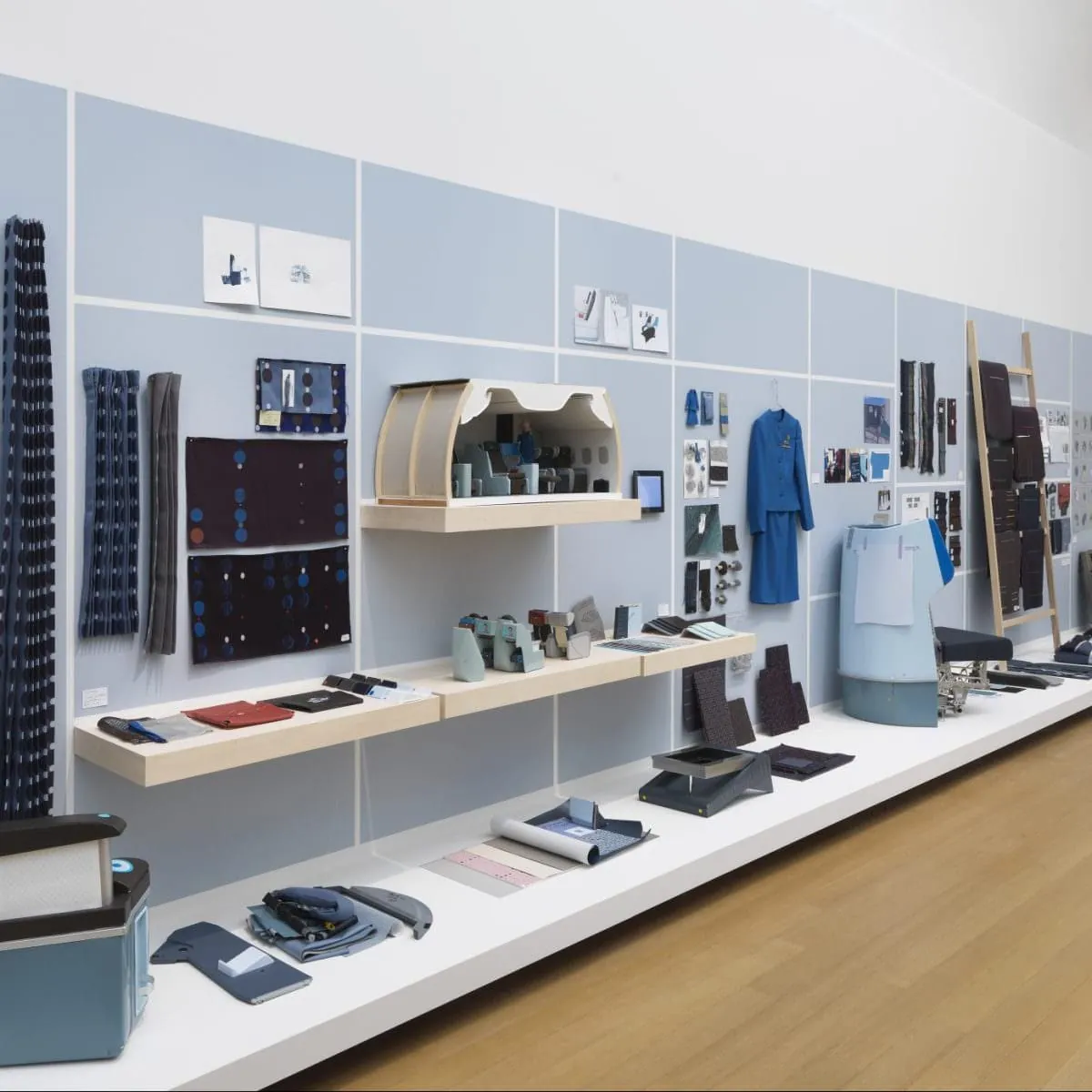 Instalación expositiva del proyecto KLM: pared azul grisácea con muestras textiles, modelo a escala de cabina con asiento, uniforme azul de azafata colgado, fragmentos de prototipo de asiento y paneles de color