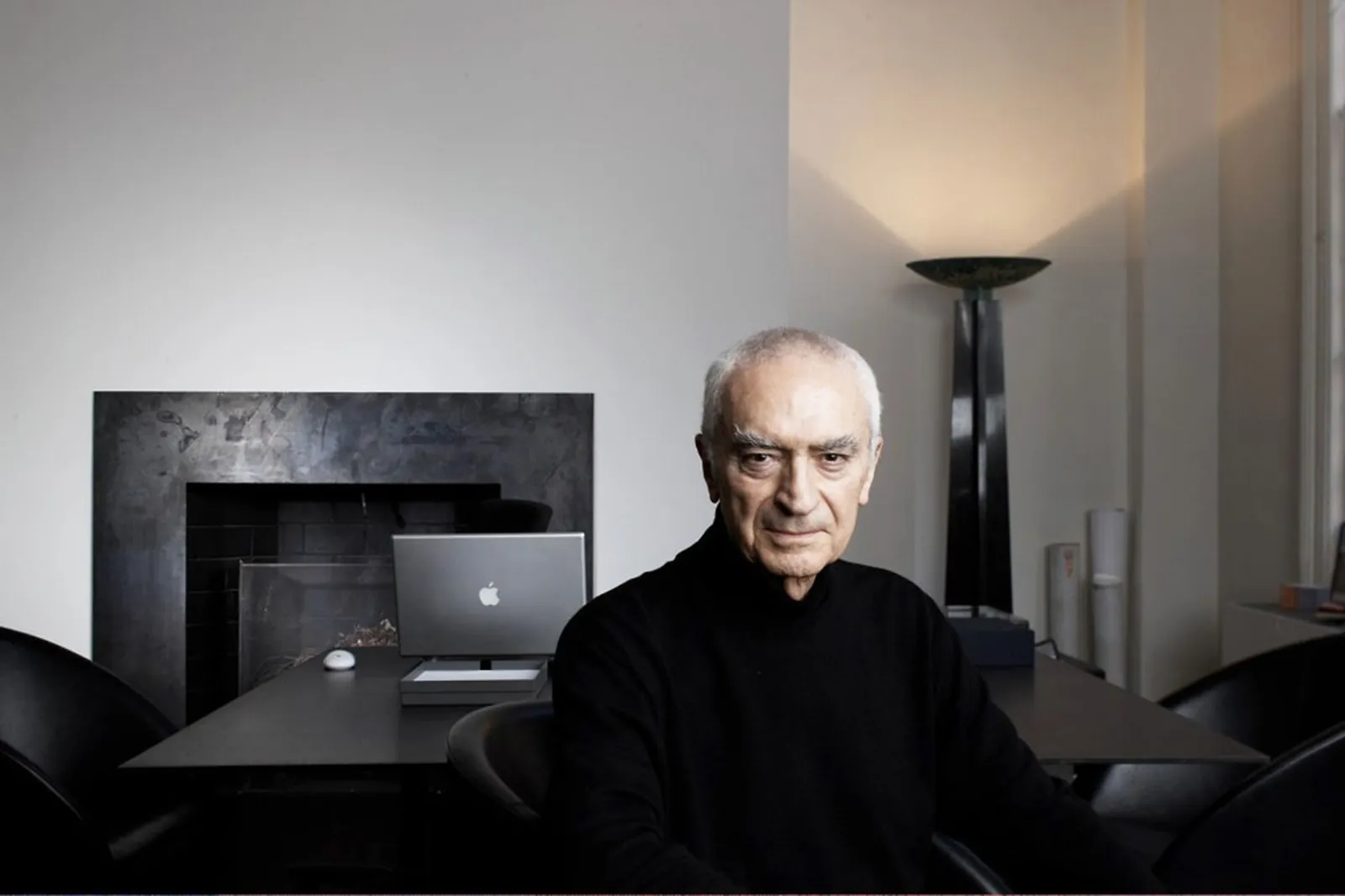 Retrato de Massimo Vignelli en su estudio con jersey de cuello alto negro, mesa con MacBook al fondo, lámpara y fireplace negro a la izquierda