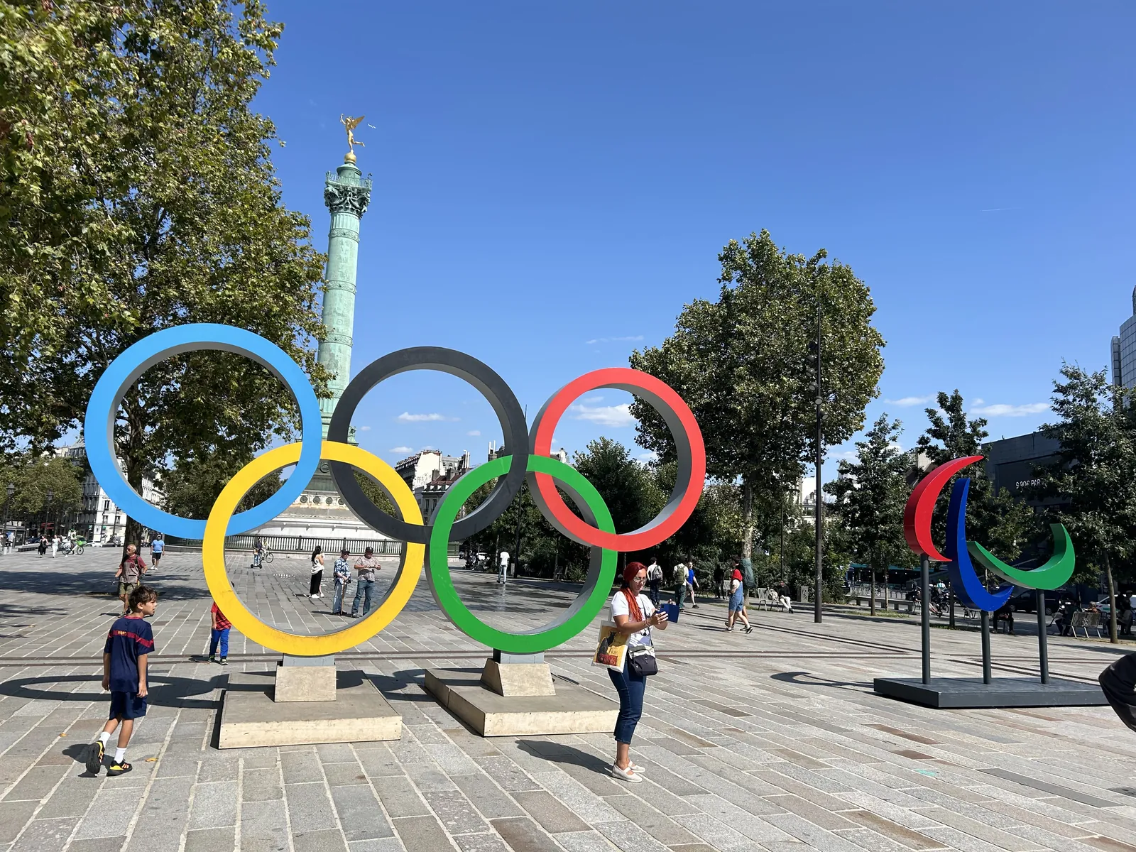Place de la Bastille en agosto de 2024 con los anillos olímpicos de gran tamaño y el agitos paralímpico instalados como esculturas urbanas, columna de Julio al fondo, transeúntes alrededor