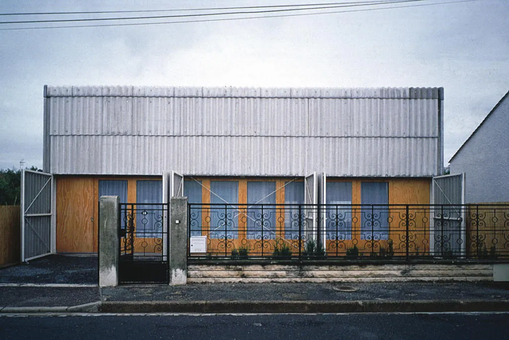 Maison Latapie en Floirac vista desde la calle: fachada con cerramiento ondulado de policarbonato translúcido en la parte superior y carpinterías de madera natural en la planta baja, valla de hierro forjado en primer plano