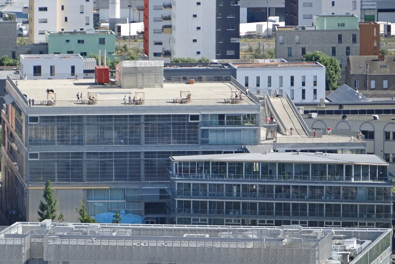 Vista cenital de la ENSA Nantes: edificio horizontal con cubierta plana habitada por personas, rampa exterior conectando niveles, contexto urbano denso al fondo