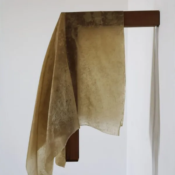 Pieza textil del Manifiesto Mediterráneo: paño de tonos terrosos colgado sobre un soporte de madera oscura, sobre pared blanca