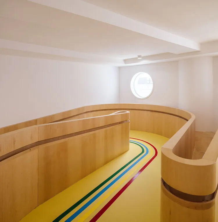 Interior de la Aceleradora La Paz: espacio doble altura con balaustrada curva de madera natural, suelo amarillo con líneas paralelas multicolor (pista deportiva infantil), ventana redonda blanca tipo ojo de buey