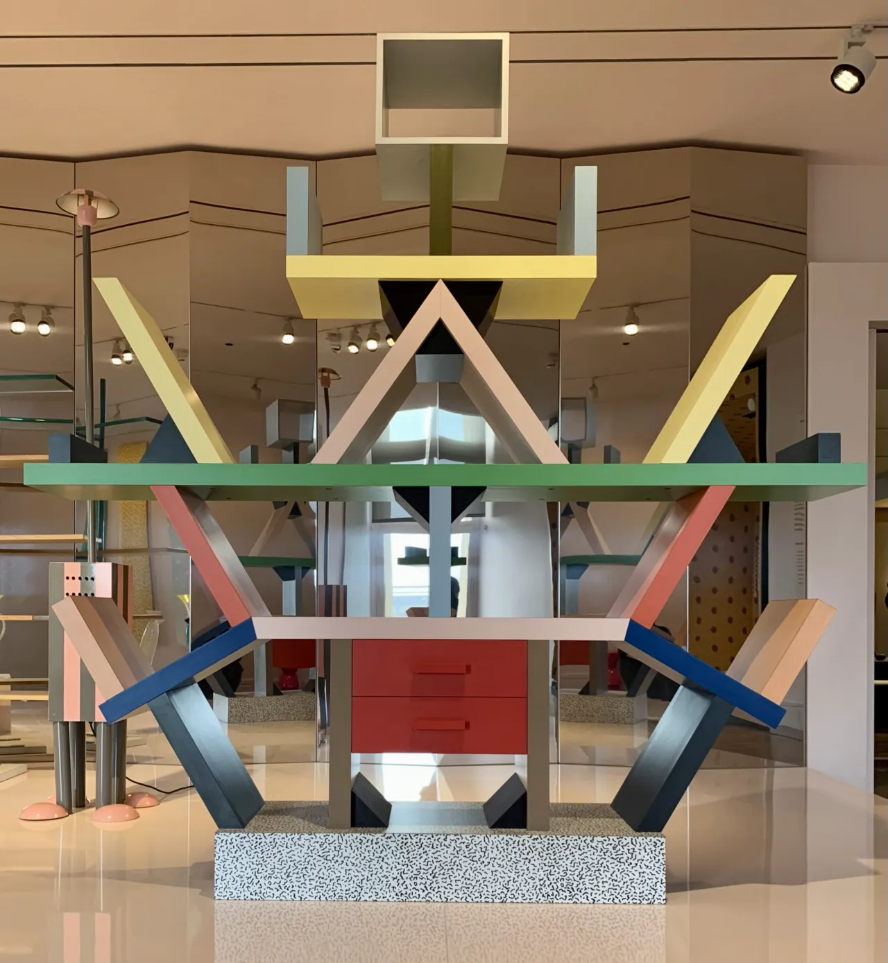 Carlton Bookcase de Ettore Sottsass para Memphis 1981: estantería de madera chapeada y laminado plástico con compartimentos asimétricos en colores chillones (verde, amarillo, rosa, azul, rojo) sobre base de terrazzo blanco con puntos negros