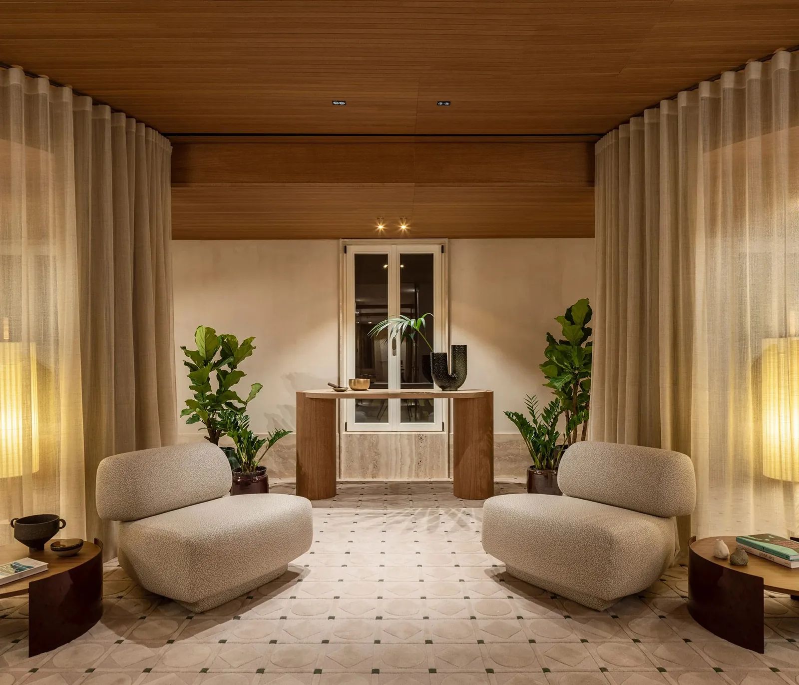 Antesala del Six Senses Rome: dos butacas blancas tapizadas en bouclé sobre suelo terrazzo, plantas en macetas, cortinas crema, lámparas de pared y consola de travertino con cerámica y planta