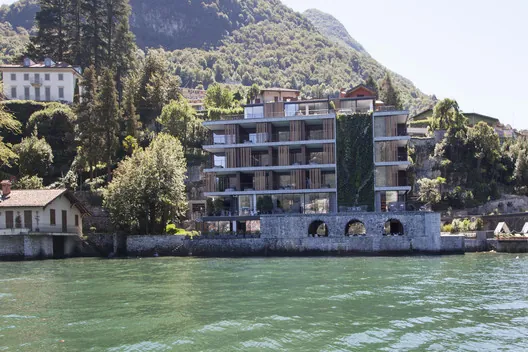 Il Sereno Hotel visto desde el Lago di Como: edificio horizontal con jardín vertical, terrazas escalonadas y arcadas en planta baja, montañas verdes al fondo
