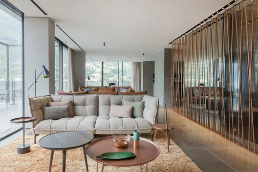 Sala de estar del Il Sereno con sofá largo gris, mesa redonda terracota y gris, ventanal al Lago di Como con montañas al fondo, divisor vertical de varas doradas a la derecha