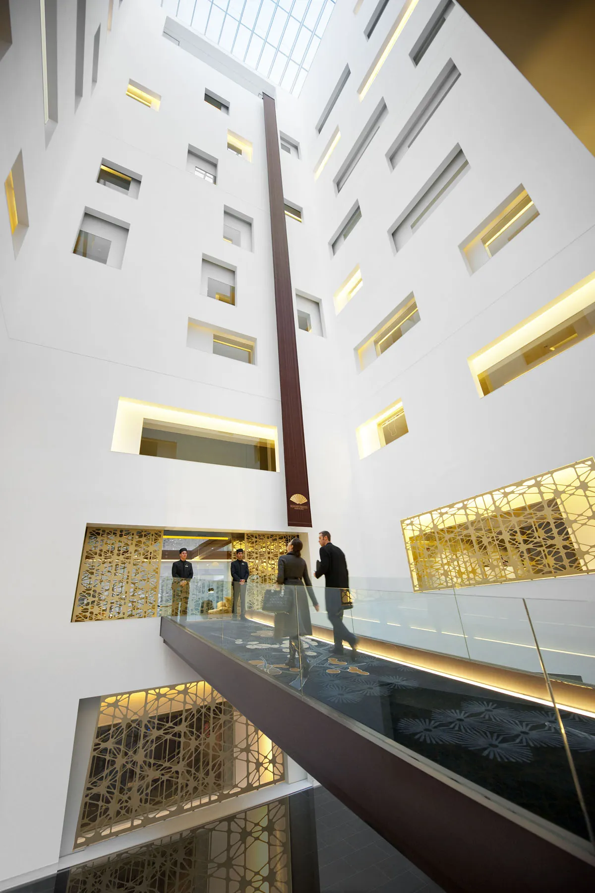 Atrio interior del Mandarin Oriental Barcelona: vacío vertical de paredes blancas con paneles dorados perforados de patrón árabe, balcón de cristal con dos personas y lucernario cenital