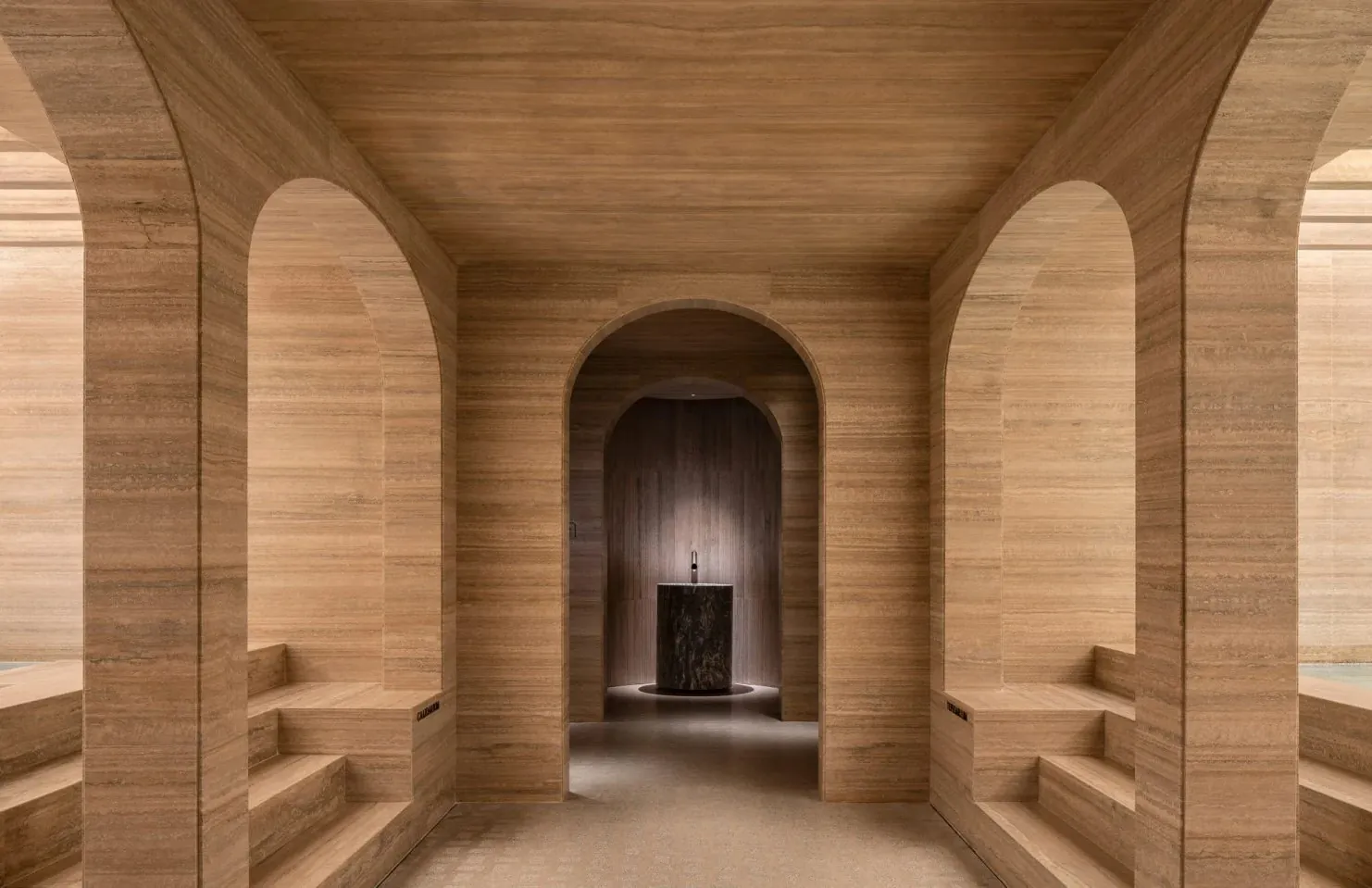 Pasillo de spa del Six Senses Rome con paredes y bóveda de travertino, arcos romanos, banco corrido de travertino, vista al fondo a una sala con fuente de mármol verde