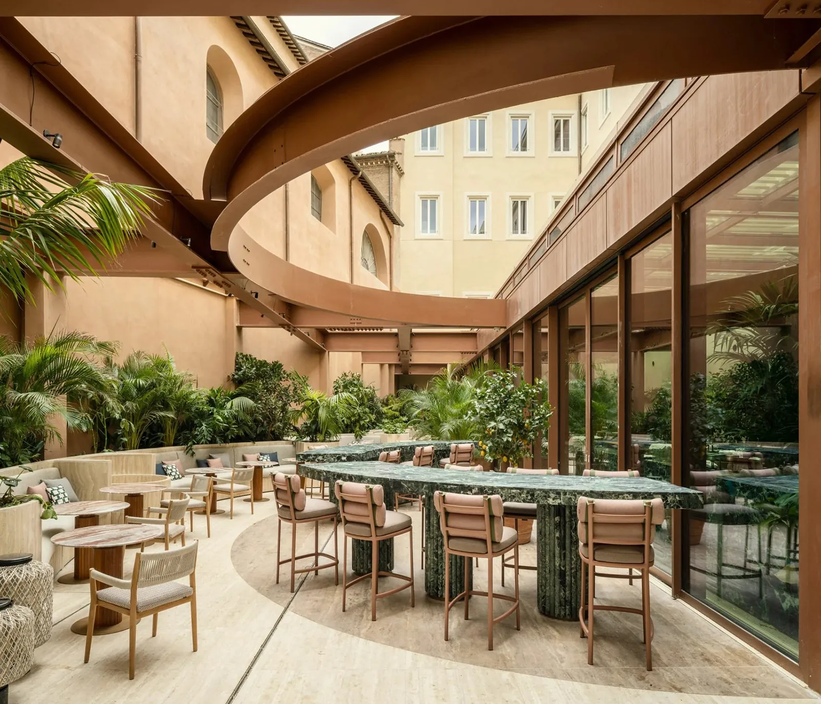 Patio interior del Six Senses Rome: mesa larga de mármol verde con sillas, cubierto por estructura curvilínea de hierro y madera que evoca balaustrada renacentista, plantas vivas en jardineras laterales, fachada del palacio renacentista al fondo