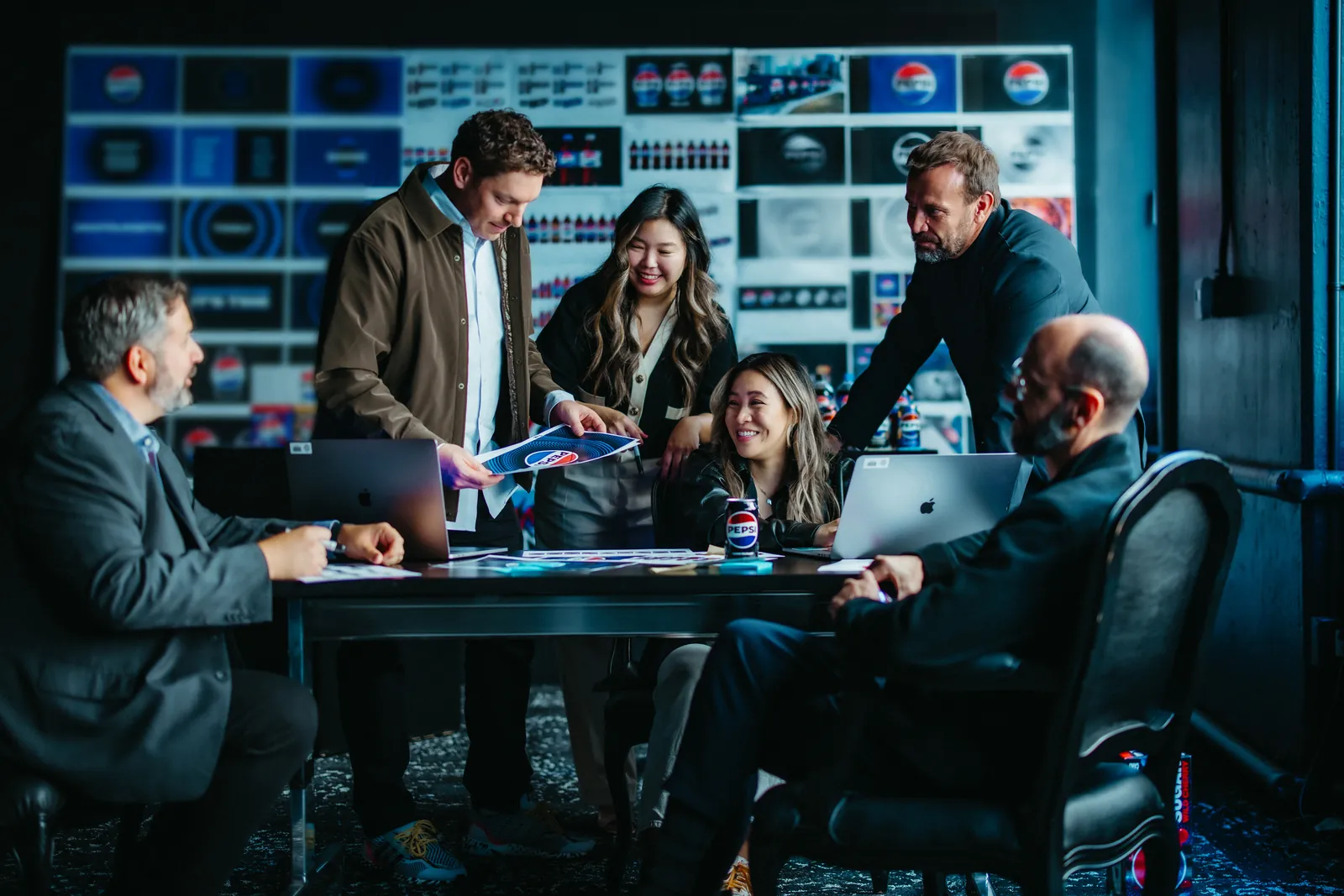 Equipo PepsiCo Design & Innovation trabajando alrededor de una mesa con laptops y latas Pepsi: cinco personas examinan piezas impresas del rebrand mientras detrás se ve un panel completo con docenas de variantes de logo exploradas
