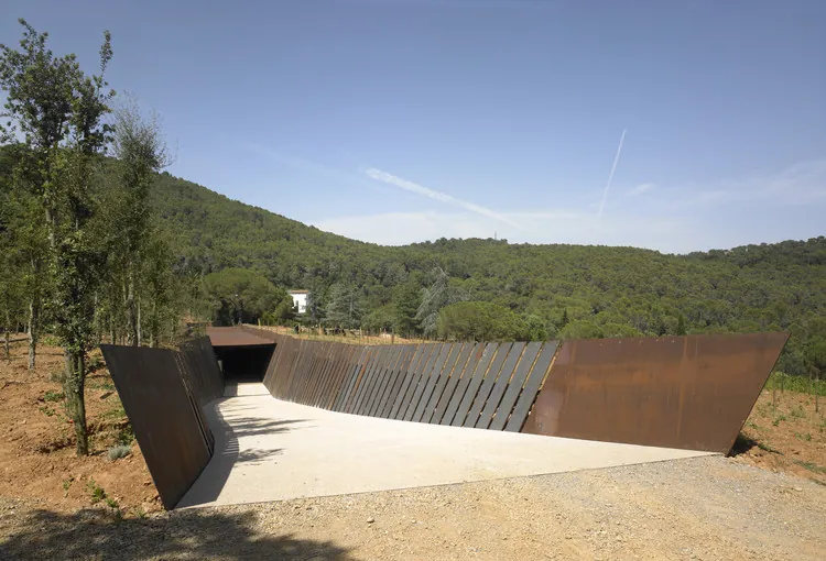 Bodega Bell-Lloc en Palamós: promenade descendente de chapas inclinadas de acero corten oxidado que articula la entrada a la bodega subterránea, vegetación catalana al fondo