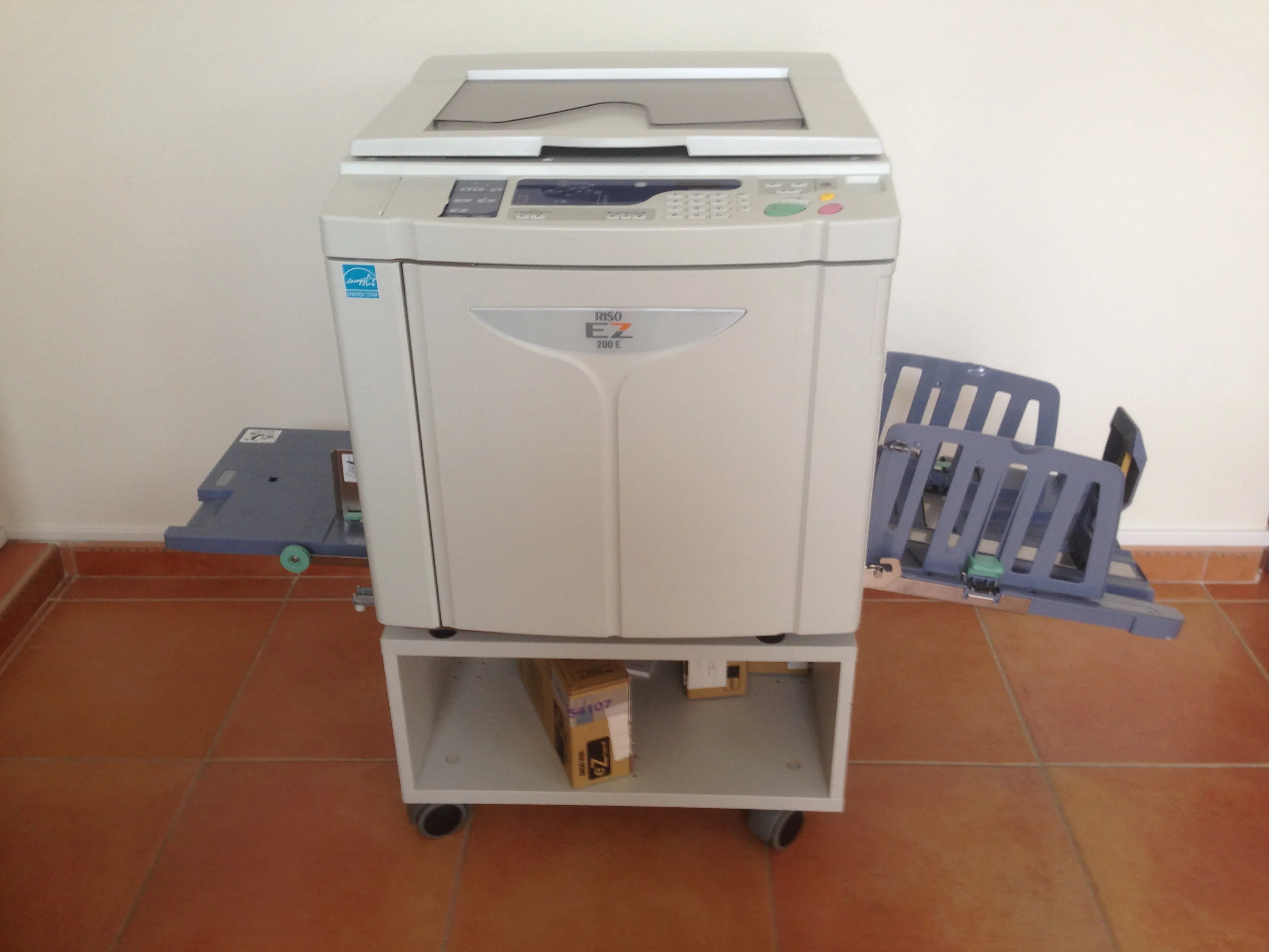 Duplicadora Riso EZ200 sobre un carrito con bandeja de salida desplegada, en una habitación con suelo de terracota — modelo compacto característico del revival riso