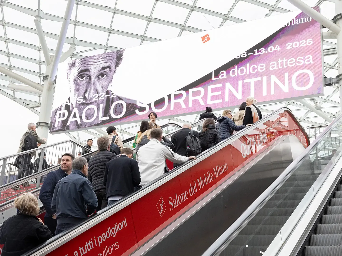 Acceso Porta Est de la Fiera Milano Rho durante el Salone 2025: escaleras mecánicas con visitantes y cartel grande de la instalación 'La dolce attesa' de Paolo Sorrentino