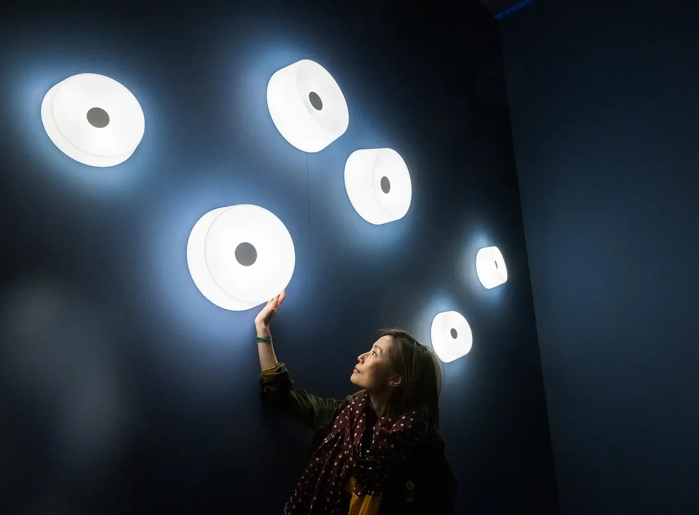 Stand de Foscarini en Euroluce 2025: instalación con seis luminarias circulares blancas suspendidas sobre fondo azul oscuro, una visitante interactúa con una de ellas