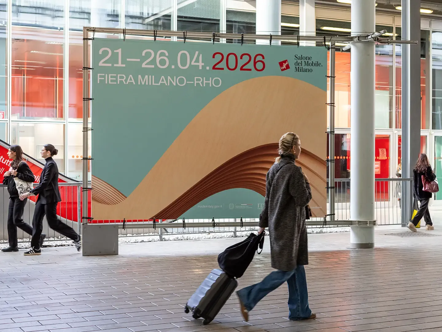Cartel grande en el recinto de Fiera Milano Rho anunciando las fechas de la próxima edición: '21-26.04 2026 Fiera Milano-Rho Salone del Mobile.Milano', con visitante de espaldas caminando con maleta