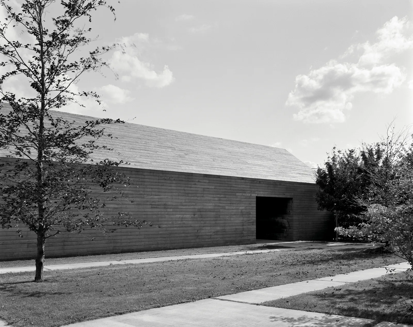 DC II Residence de Vincent Van Duysen: granja belga reconvertida, fachada de madera horizontal con cubierta a dos aguas, jardín con árboles jóvenes en primer plano, fotografiada por Hélène Binet