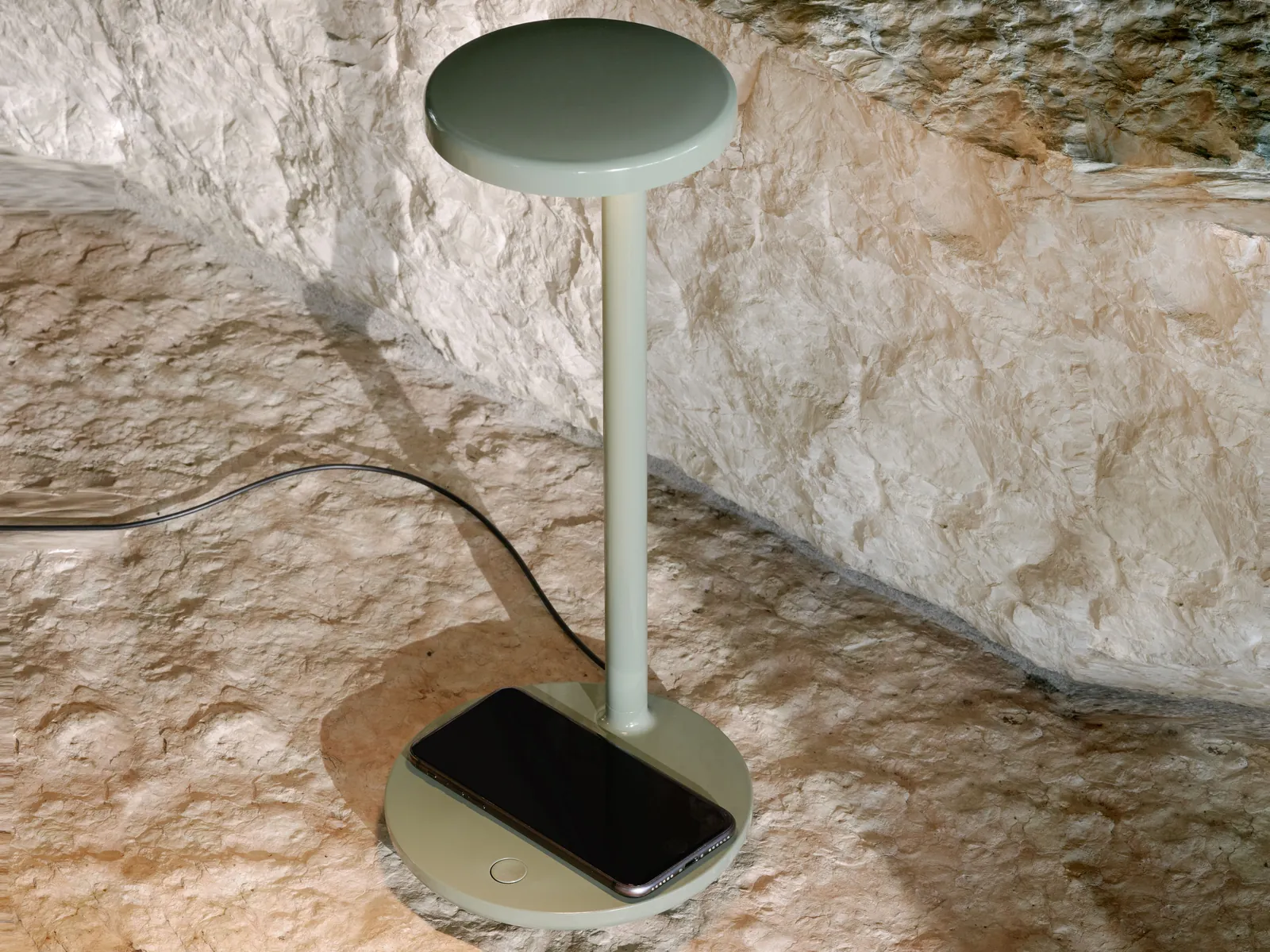 Lámpara de mesa Oblique de Vincent Van Duysen para Flos en acabado salvia, con un teléfono móvil sobre la base de carga inalámbrica, fondo de pared rugosa de piedra natural