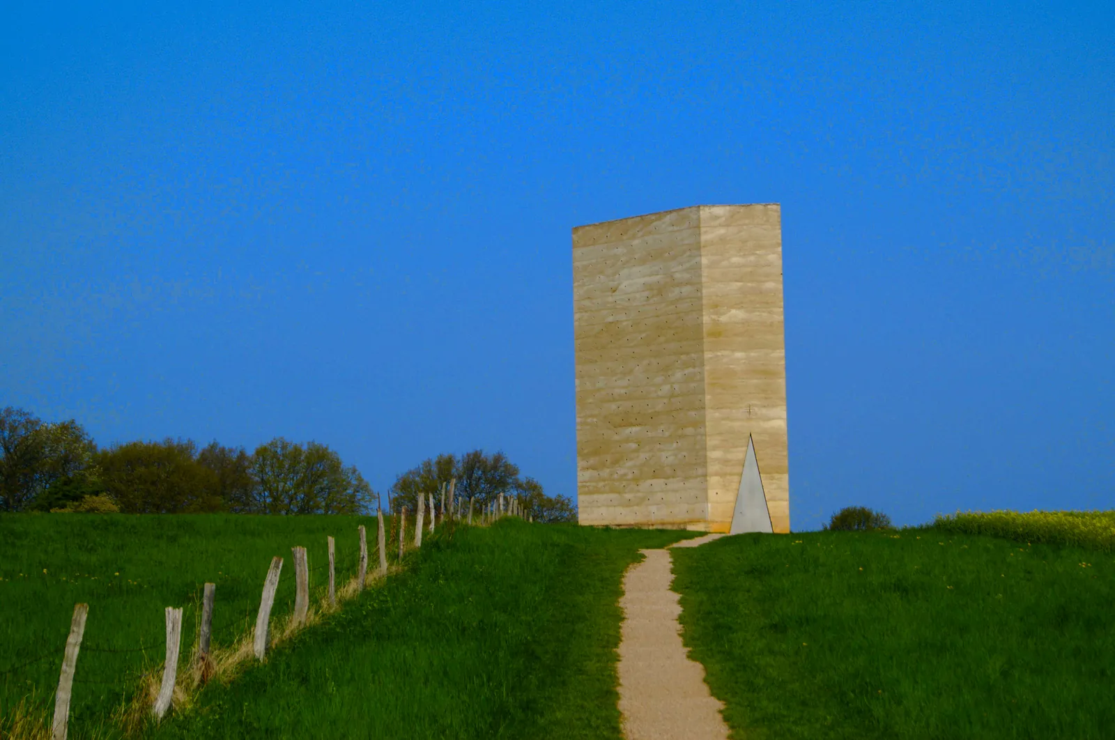 Capilla Bruder Klaus de Peter Zumthor en Wachendorf: torre rectangular de hormigón color arena con triángulo de acceso, alzada sobre prado verde bajo cielo azul intenso